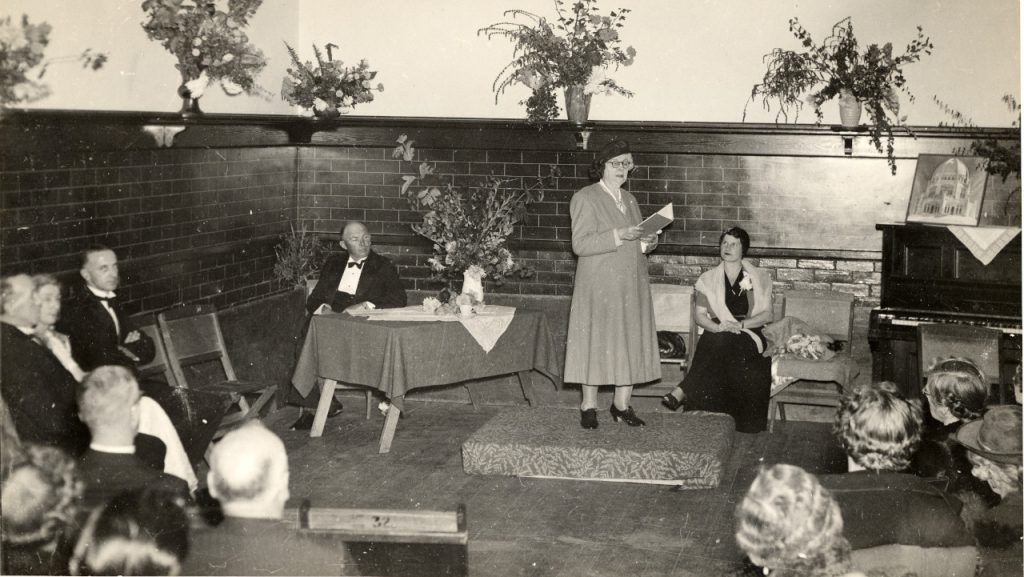 People in formal attire seated around the room watching Clara Dunn reading from a small book