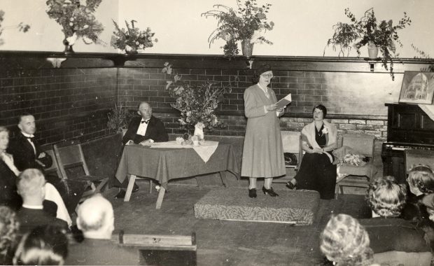People in formal attire seated around the room watching Clara Dunn reading from a small book