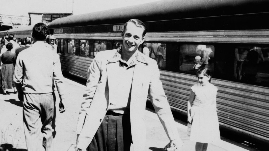 Smiling man holding suitcase in front of train with passengers behind him